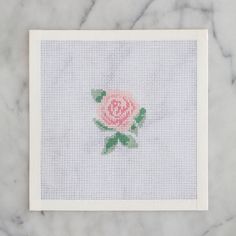 a cross - stitch rose on a white card with green leaves in the middle, sitting on a marble surface