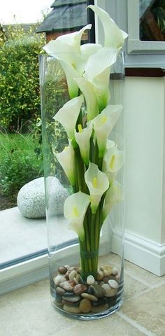 white flowers in a glass vase with rocks and pebbles on the ground near a window