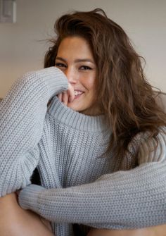 a woman with long brown hair wearing a gray sweater and smiling at the camera while sitting on a couch