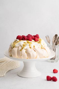 a white cake with raspberries on top sitting on a plate next to silverware