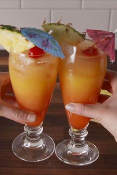 two people holding up glasses with drinks in them on a wooden counter top, one is filled with fruit and the other has an umbrella