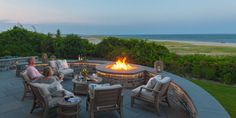 two people sitting around a fire pit with chairs and tables on the patio overlooking the ocean