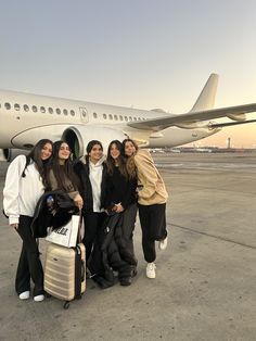 four women pose with their luggage in front of an airplane on the tarmac at sunset