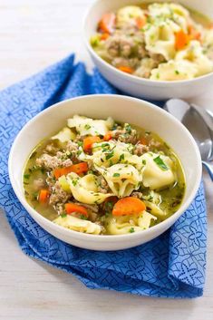 two bowls filled with pasta and meat soup on top of a blue napkin next to silver spoons