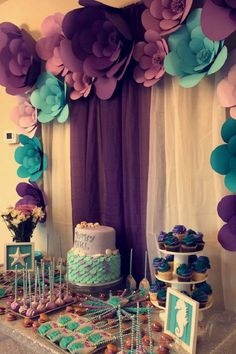 a table topped with cake and cupcakes covered in purple and blue paper flowers