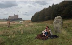 two people sitting on the grass in front of a castle with horses grazing behind them