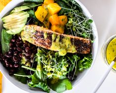 a white bowl filled with lots of food next to a glass of olive dressing on top of a table