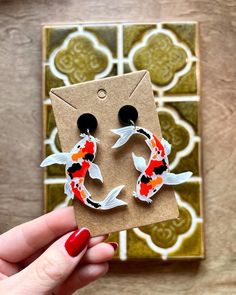 a pair of handmade koi fish earrings on top of a cardboard card next to a tile wall