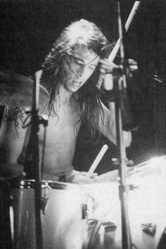 a man with long hair playing drums in a black and white photo, while looking down at the drum sticks