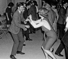 an old photo of two people dancing in a room with other people standing and sitting around