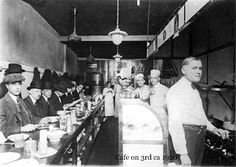 an old photo of men in hats at a bar with food on the counter and one man standing next to him