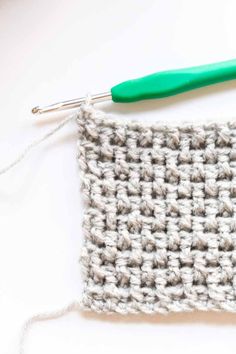 the crochet square is being worked on with a green handled knitting needle in front of it