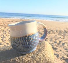 a coffee cup sitting on top of a rock at the beach in front of the ocean