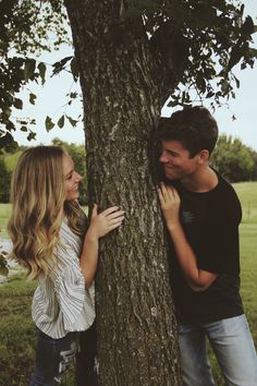 a man and woman standing next to each other near a tree in the park, hugging