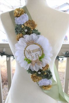 a white mannequin wearing a green sash with flowers and the words mommy to be written on it