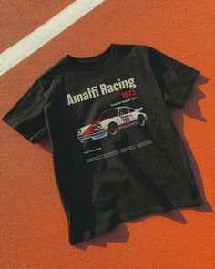 Step into the racing history of Sicily with our Amalfi Racing 1973 Tee. Inspired by the Targa Florio, this tee brings the excitement of coastal speed and vintage elegance to your wardrobe. Unisex soft cotton crew neck t-shirt featuring a Amalfi Racing Vintage Car graphic printed on the front. Fabrics: 100% cotton, 6 oz Made in: LA Casual Short Sleeve T-shirt For Motorsport Events, Racing Style T-shirt With Letter Print And Crew Neck, Racing Style Crew Neck T-shirt For Motorsport Events, Black Graphic Print Tops For Motorsport Events, Casual T-shirt With Letter Print For Motorsport Events, Cotton T-shirt For Motorsport Events, Cotton Short Sleeve T-shirt For Motorsport Events, Cotton Racing Style Top With Graphic Print, Casual Crew Neck Tops For Motorsport Events