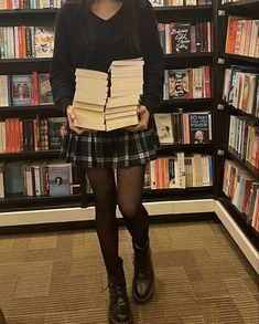 a woman holding several books in front of a book shelf filled with books and wearing black boots