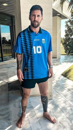 a man standing in front of a building with tattoos on his arms and legs, wearing a blue soccer jersey