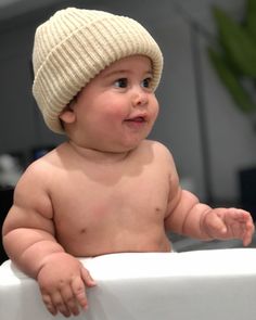 a baby wearing a knitted hat sitting in a bathtub looking at the camera