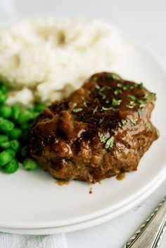 a white plate topped with meat covered in gravy next to peas and mashed potatoes