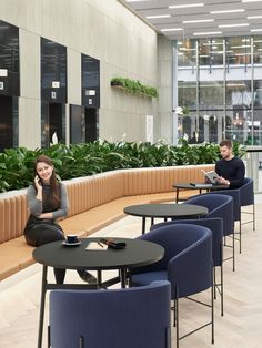 two people sitting at round tables in an office building with plants on the wall and windows