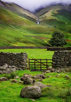 a stone wall and gate in the middle of a green field with mountains behind it