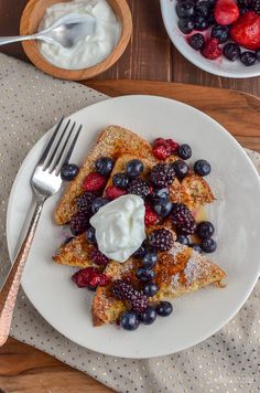 french toast with berries, blueberries and whipped cream on a plate next to a bowl of yogurt