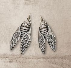 two silver earrings with wings hanging from them on a wooden surface in front of a concrete wall