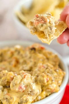 a hand holding up a cracker over a bowl of dip with cheese and meatballs