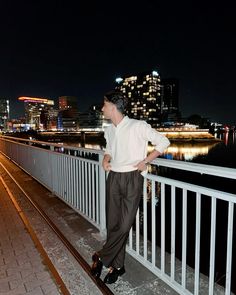 a man is standing on a bridge looking at the city lights in the distance with his hands on his hips