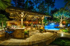 an outdoor kitchen and dining area next to a swimming pool at night with lights on