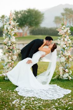 a bride and groom kissing in front of an arch with flowers on the grass at their wedding