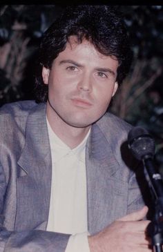 a man sitting in front of a microphone with his hand on the microphone and looking at the camera