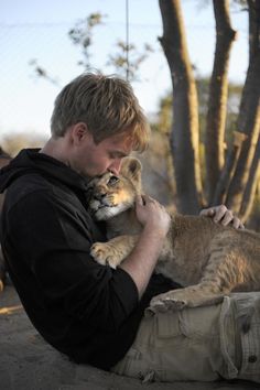 a man holding a small lion cub in his lap while sitting on the ground next to a tree