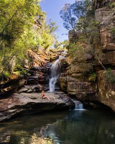 a person sitting on the edge of a waterfall next to a body of water and trees