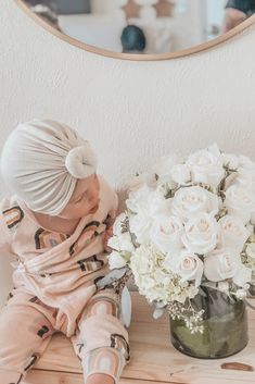 a baby sitting on a table next to a vase filled with white flowers and a mirror