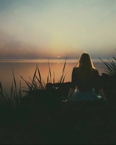 a woman sitting on top of a grass covered hillside next to the ocean at sunset