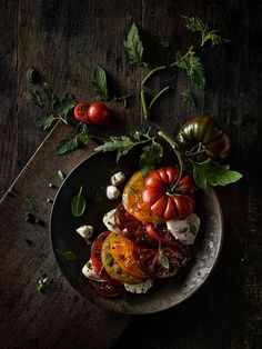 a black plate topped with tomatoes and other vegetables on top of a wooden table next to green leaves