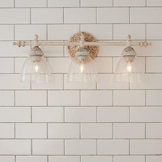 three lights are on the wall in front of a white tiled wall and below them is a light fixture