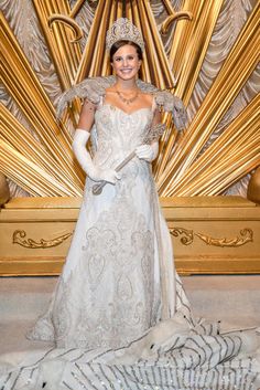 a woman wearing a tiara and gloves standing in front of a golden wall with feathers