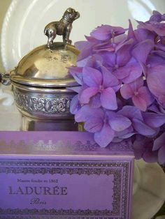 purple flowers are sitting on a table next to a silver container with a card in it