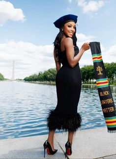 a woman in a black dress and hat is holding a scarf by the water's edge