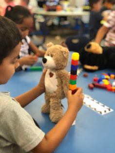 a young boy holding a teddy bear in front of other children sitting at a table