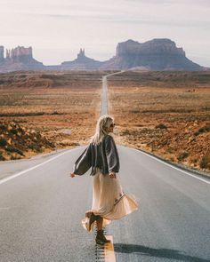 a woman is walking down the middle of an empty road with mountains in the background