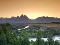 the sun is setting over some mountains and trees with a river running through it in the foreground