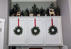 Christmas wreaths hanging on ribbon attached to kitchen cabinet doors. Different sizes of fake green Christmas trees on cabinet top with wooden reindeer for a refined but rustic holiday display. Cabinet Top Christmas Decorating Ideas, Garland On Top Of Cabinets, Upper Cabinet Christmas Decor, Garland Above Cabinets, Above Cupboard Christmas Decor, Above The Cabinet Christmas Decor, Christmas Decorations Above Cabinets, Present Cabinets Christmas, Christmas Decor On Top Of Cabinets