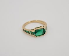 At the centerpiece of this lab grown Zambian emerald ring is a radiant 1.5 ct emerald, set east west to highlight its natural allure. Flanking it are calibre cut emeralds, each uniquely shaped to create a seamless gradient effect, making every ring truly one-of-a-kind. Set in luxurious 18K yellow gold, this ring exudes timeless charm. What makes it truly special? Calibre cut stones are individually shaped to fit precisely into the design, resulting in a mesmerizing display of color and brillianc Formal Green Channel Set Emerald Ring, Timeless Baguette Cut Green Emerald Ring, Green Emerald-cut Channel Set Jewelry, Modern Green Emerald Ring With Accent Stones, Timeless Green Baguette Cut Emerald Ring, Timeless Green Baguette-cut Emerald Ring, Emerald Channel Set Jewelry With Emerald Cut, Emerald Cut Channel Set Emerald Jewelry, Modern Octagon Emerald Ring