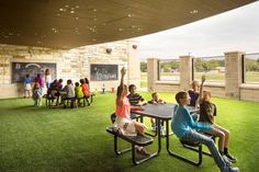 children are sitting at picnic tables on the grass in front of a wall with chalkboards