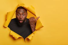 a man making a face through a hole in yellow paper with his finger pointing at the camera