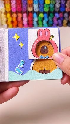 a person holding up a small card with a bear on it and colorful crayons in the background